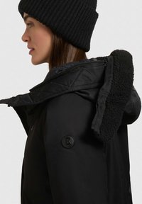 Jachetă neagră cu glugă, având o margine interioară moale din fleece și un material exterior neted. Include o mică etichetă logo neagră pe mânecă.