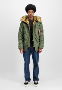 Green padded jacket with a faux fur hood, orange lining, and multiple buttons. Paired with dark blue jeans and black sneakers.