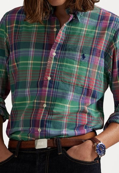 Homme portant une chemise à carreaux verte, violette et rouge, rentrée dans un jean foncé avec une ceinture en cuir marron et une montre, les mains dans les poches.
