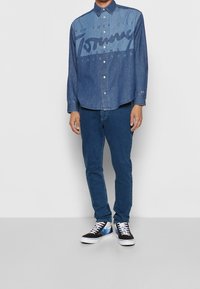 Camisa de denim con logo frontal en azul oscuro, diseño de botón, combinada con jeans ajustados en azul oscuro y zapatillas negras con un patrón colorido.
