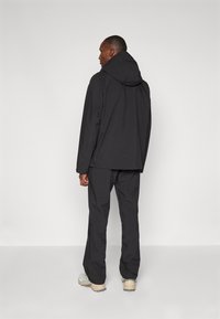 Veste imperméable noire avec capuche, associée à un pantalon noir assorti. Texture lisse, coupe ample et design minimaliste. Chaussures de couleur claire.