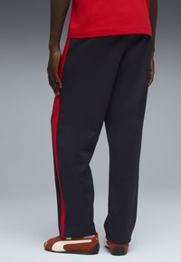 Σκούρο μπλε φόρμα με κόκκινες ρίγες στο πλάι, διαθέτει άνετη εφαρμογή, ομαλό ύφασμα και ελαστική μέση. Συνδυασμένο με καφέ και λευκά αθλητικά παπούτσια.