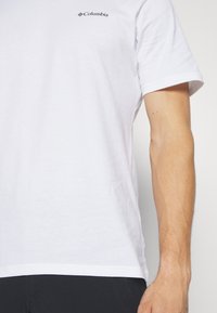 T-shirt en coton blanc à manches courtes, avec un petit logo noir de Columbia sur le côté gauche de la poitrine et un ourlet droit.