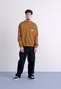 Sudadera marrón con un parche de logo en el pecho, combinada con pantalones negros de corte relajado y zapatillas negras sobre un suelo azul.