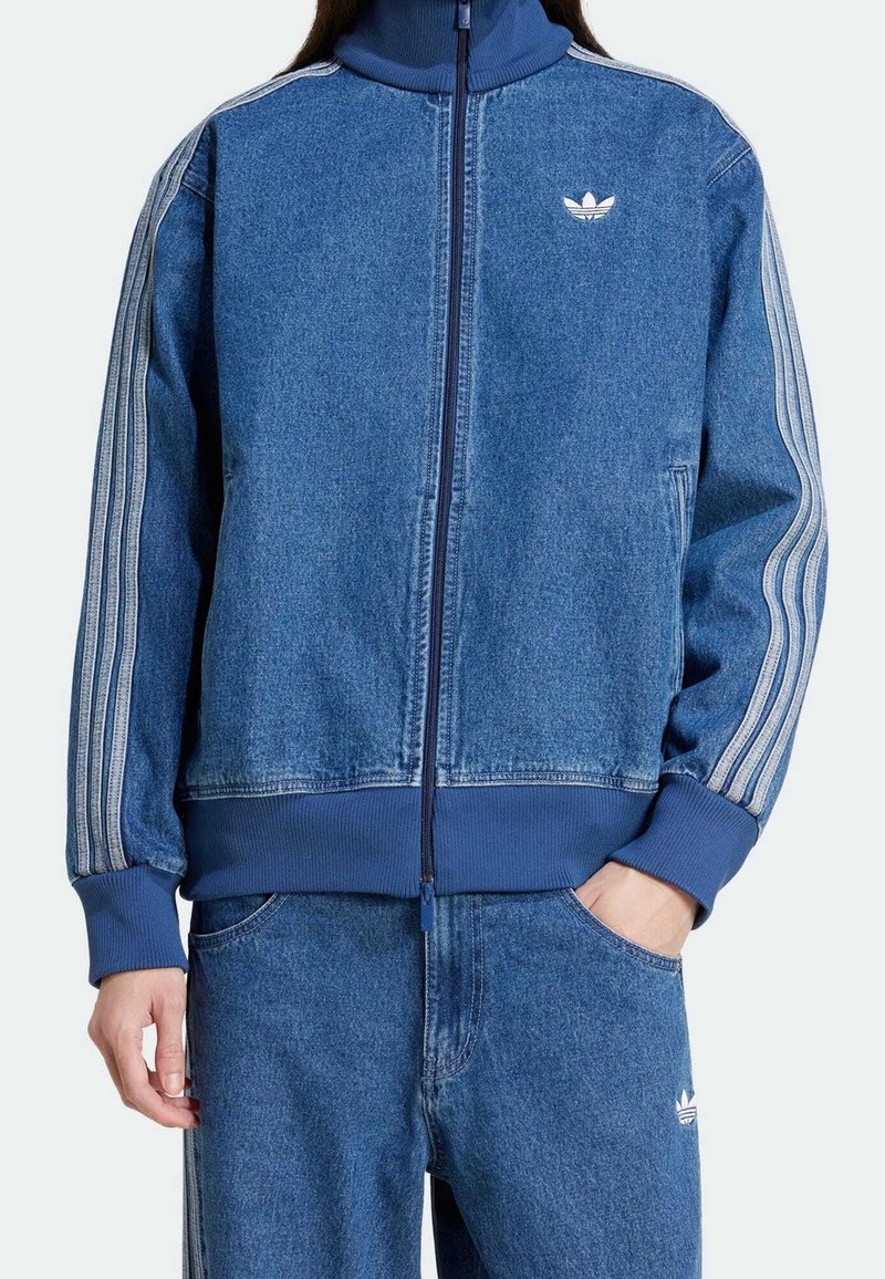 Chaqueta de mezclilla azul con cremallera y cuello alto, puños de canalé y tres rayas blancas a lo largo de las mangas. Presenta un logo bordado en el pecho y bolsillos.