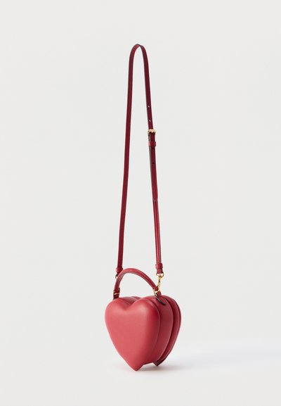 Sac à main en cuir rouge en forme de cœur avec une longue bandoulière réglable et une petite poignée, présenté sur un fond clair uni.