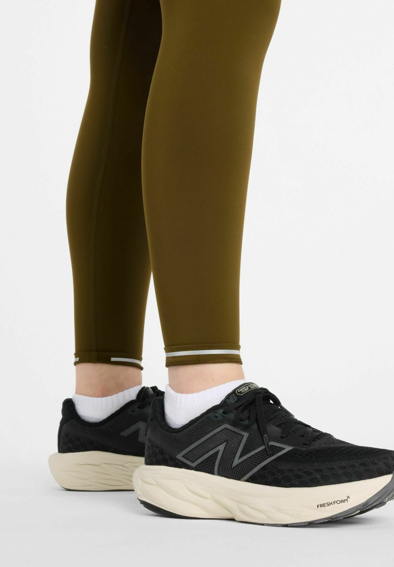 Svarta träningsskor med texturerad ovandel och krämfärgad sula. Olivgröna leggings med slät finish och vita accentstripes vid mudden.