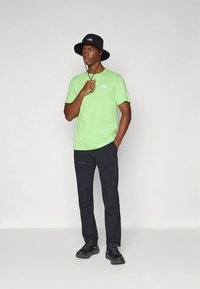 Grünes kurzärmeliges Hemd, schwarze Hose und schwarzer Fischerhut. Das Hemd verfügt über ein kleines Logo, die Hose hat eine schlanke Passform und dezente Akzente. Schwarze Schuhe.