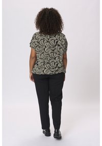 Frau mit lockigem Haar, die ein kurzärmeliges Top mit schwarz-beigem Muster, schwarze Hosen und schwarze Schuhe trägt und mit dem Rücken zu einer einfarbigen Hintergrund steht.