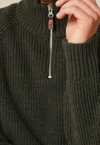 Pull en maille de couleur vert foncé avec un col montant et une demi-fermeture éclair. Le tissu texturé présente un motif côtelé et un accent de tirette en cuir.