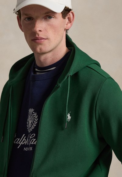 Noor mees kannab tumerohelist lukuga kapuutsi sinise Ralph Laureni dressipluusi ja valget nokkmütsi, taust on neutraalne.