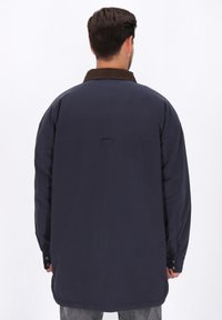 Ein Mann trägt eine dunkelblaue, oversized Jacke mit einem braunen Cordkragen, von hinten gegen einen schlichten weißen Hintergrund gezeigt.