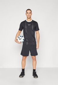 Zwarte sportieve t-shirt met verticale strepen, witte kraag, logo op de borst. Gecombineerd met zwarte shorts. Model houdt een voetbal vast.