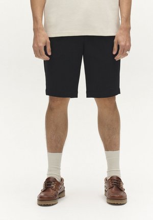 Osoba oblečená v čiernych šortkách, béžových ponožkách, hnedých kožených topánkach a béžovom textúrovanom topu, stojaca pred bielym pozadím.