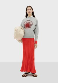 Sweatshirt gris avec un graphique de soleil rouge, associé à une jupe maxi plissée rouge. Le modèle tient un sac beige et porte des sandales en fourrure marron.