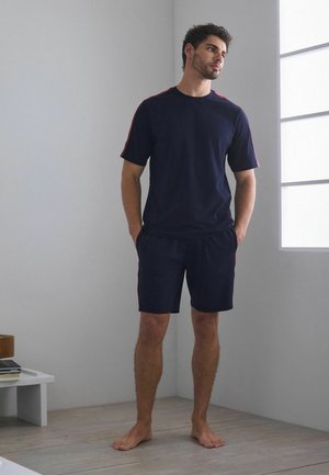 Jonge man staat op blote voeten in een lichte kamer, draagt een marineblauw T-shirt en een korte broek met rode strepen, handen in de zakken, kijkt opzij.