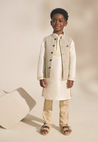 L'ensemble traditionnel pour un enfant se compose d'un kurta crème, d'un gilet gris avec des boutons, de pantalons beiges et de sandales brunes avec des boucles.