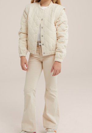 Persoon draagt een gebroken-witte doorgestikte jas over een wit shirt, beige uitlopende broek en witte sneakers, staand tegen een neutrale achtergrond.