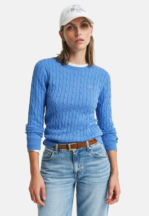 Pull en maille torsadée bleu clair avec des poignets et une taille côtelés, associé à un jean bleu délavé et une casquette de baseball blanche avec du texte.