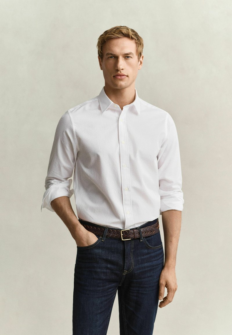 Blond man with folded sleeves white shirt, dark jeans, and brown braided belt stands with left hand in pocket on beige background.