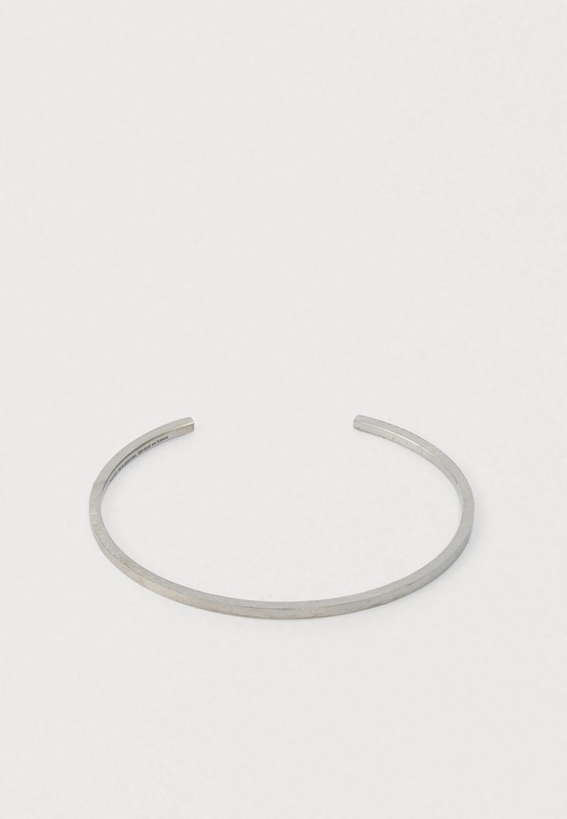 Plonas sidabrinis atviras apyrankė su lygiu, šiek tiek tekstūruotu paviršiumi, padėta ant vientiso balto fono.