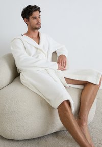 Accappatoio bianco in spugna con cappuccio, caratterizzato da maniche lunghe e una vestibilità comoda. Indossato mentre si è seduti su una sedia a sdraio leggera e strutturata.