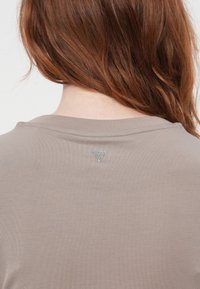 Žena s vlasy po ramena v barvě červené, oblečená v taupe topu s malou stříbrnou trojúhelníkovou dekorací na zadním výstřihu.
