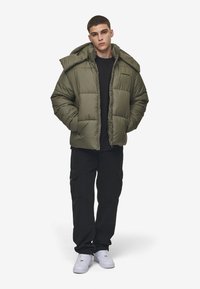 Olive green puffer jacket with a zip front, large hood, and quilted design, paired with black cargo pants and white sneakers.