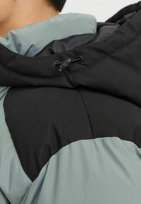 Padded hooded jacket featuring a black top section and a light green lower section, made of textured fabric with adjustable drawstrings.