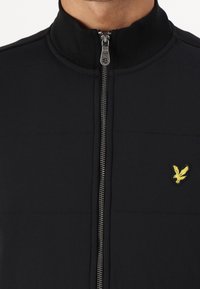 Giacca nera con zip e colletto alto, realizzata in tessuto testurizzato, con logo dell'aquila giallo a contrasto e dettagli della zip argentati.