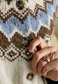 Przypinanie guzika na wełnianym swetrze z dzianym wzorem w zygzaki w odcieniach brązu, niebieskiego i białego, na palcu złoty trójkątny pierścionek.