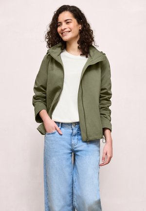 Smilende kvinde med krøllet hår iført en olivengrøn jakke, hvid skjorte og lyseblå jeans med den ene hånd i lommen.