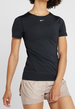 Vrouw in een nauwsluitend zwart Nike sportshirt met korte mouwen en beige gelaagde hardloopshorts, staand tegen een effen achtergrond.
