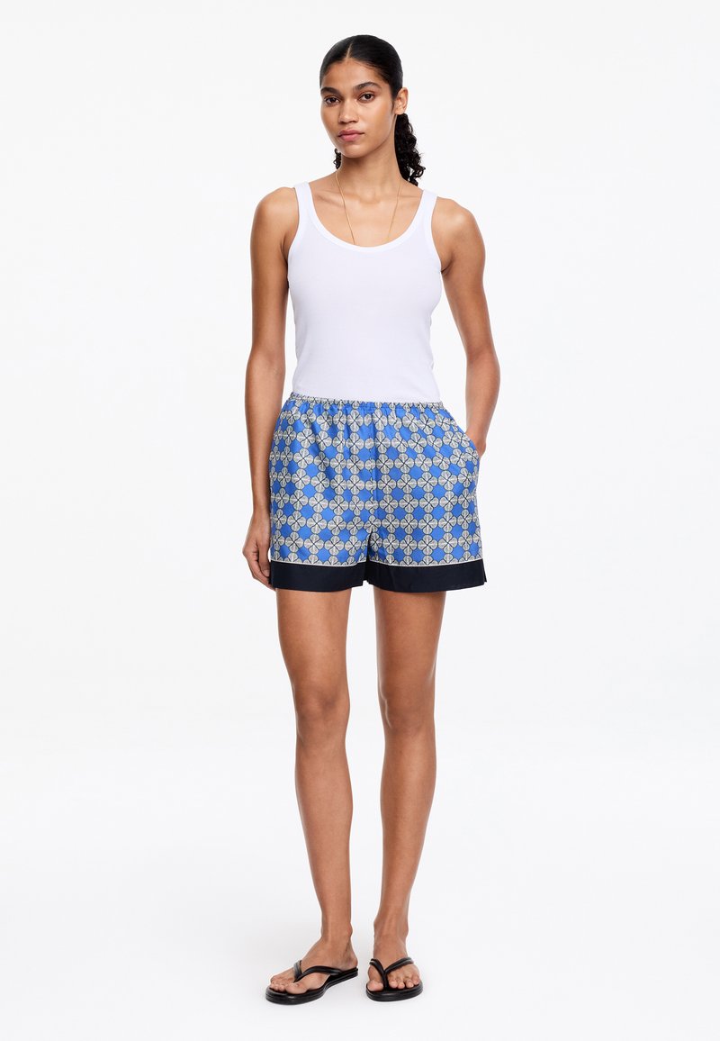 Femme debout portant un débardeur blanc, un short bleu à motifs avec un bord noir et des tongs noires, mains dans les poches, sur fond blanc.