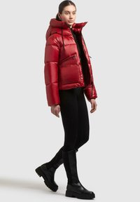 Vrouw in een glanzende rode pufferjack, zwarte leggings en zwarte laarzen tot de knie staat met één voet iets omhoog.