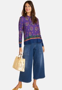 Purple patterned blouse featuring floral designs, paired with wide-leg denim jeans. Holds a gold metallic handbag and wears matching shoes.