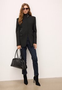 Sort skræddersyet blazer med strukturerede skuldre, parret med mørke denim jeans. Modellen holder en sort læderhåndtaske og har sorte højhælede sko på.