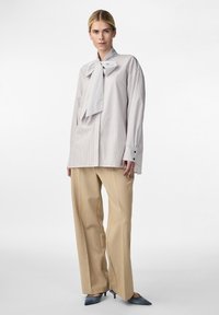 Chemise rayée blanche et grise avec un grand col en nœud, associée à un pantalon large beige et des escarpins bleus à bout pointu.