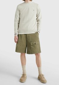 Suéter de punto gris claro con mangas largas y escote redondo; pantalones cortos de cargador verde oliva con bolsillos frontales; zapatos y calcetines beige.
