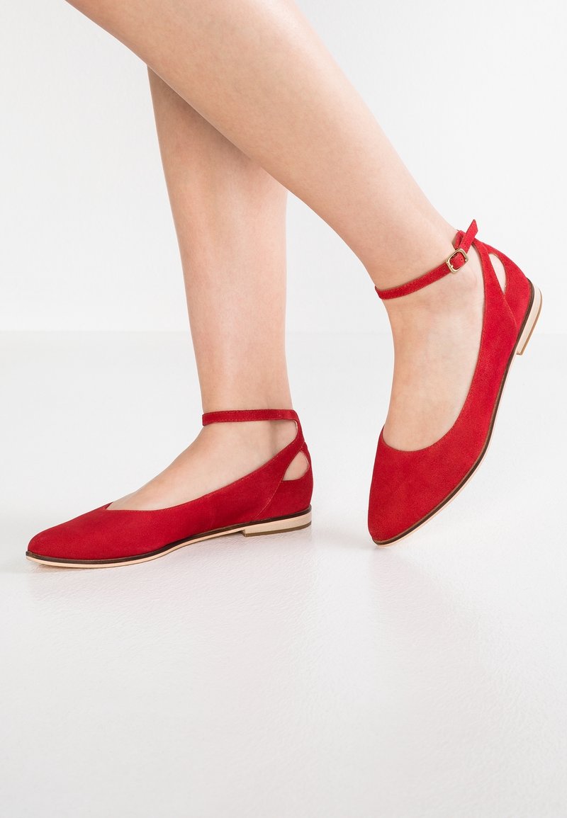 Jambes portant des chaussures plates en daim rouge avec des brides de cheville et des boucles dorées, posées sur un sol et un fond blancs.