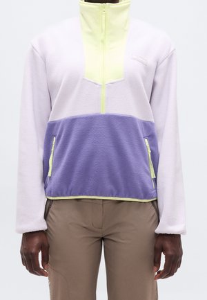 Personne portant une veste polaire bicolore pastel avec une fermeture éclair jaune et des poches, associée à un pantalon beige.