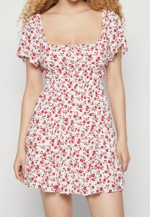 Kvinde iført en hvid kjole med rødt blomsterprint, korte pufærmer, rynket talje og bundet halsudskæring.