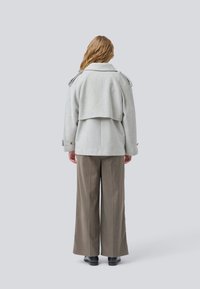 Henkilö, jolla on aaltoileva hius, kääntyy poispäin, pukeutuneena vaaleanharmaaseen vyöllä varustettuun takkiin, jossa on olkaremmejä, leveälahkeisiin ruskeisiin housuihin ja mustiin kenkiin.