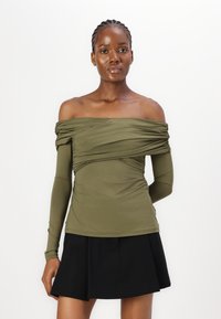 Olivu zaļš off-shoulder tops ar garām pieguļošām piedurknēm, savilktas auduma drapēšanas un gluda tekstūra, kopā ar melnu krokoto svārku.