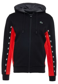 Felpa nera con zip, pannelli laterali rossi, lacci neri e loghi di coccodrillo bianchi lungo le maniche e il petto.