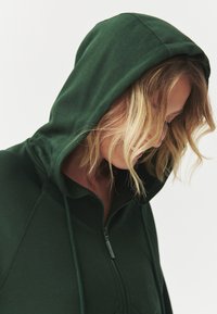 Sudadera con cremallera de color verde oscuro, con capucha ajustable y mangas raglán, que presenta una textura de tela suave. Vista cercana de la zona del cuello.