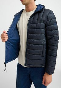 Marineblå puffjakke med horisontal quiltet tekstur, glidelås foran og hette. Foret interiør med to sidelommer og justerbar snor i bunnen.