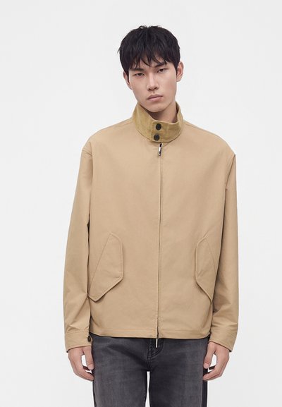 Jeune homme portant une veste beige zippée avec un col montant et deux poches avant, associée à un pantalon gris foncé, debout sur un fond uni.