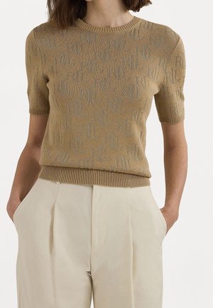 Femme portant un haut en maille beige à manches courtes avec des lettres subtilement imprimées et un pantalon plissé crème taille haute, les mains dans les poches.