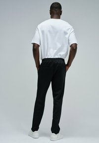 Camiseta blanca de algodón y pantalones de chándal negros con cintura elástica. Los pantalones cuentan con bolsillos laterales. El modelo lleva zapatillas deportivas blancas.